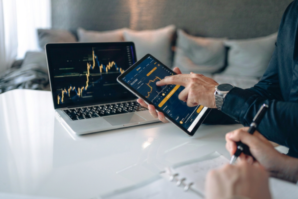Person uses a tablet displaying a stock trading app while a laptop beside shows a market chart on a white desk, with a notebook in view.