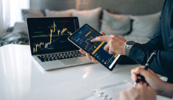 Person uses a tablet displaying a stock trading app while a laptop beside shows a market chart on a white desk, with a notebook in view.